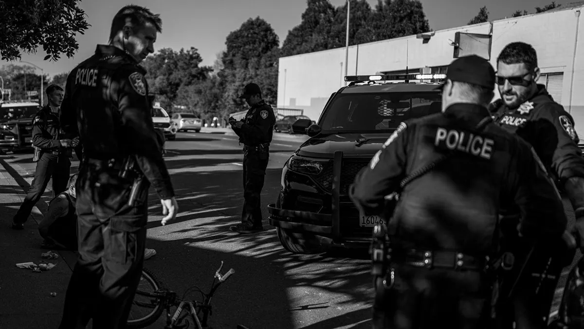 Several Vallejo police officers stand around an unmarked patrol SUV on a city street. A man sits on the ground near the officers, and a bicycle lies on its side on the pavement nearby.